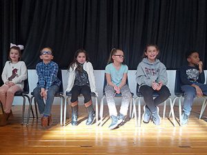 picture of students sitting on chairs on elementary school stage before spelling bee