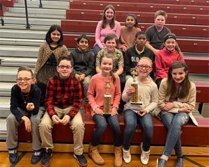 picture of the 13 contestants who competed in the spelling bee seated on the bleachers smiling