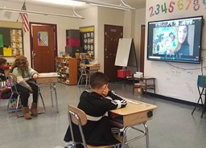 students seated as desks listen to a story read by virtual presenter on screen to prepare for ice cream making activity