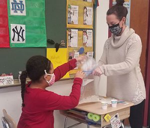 a teacher holds a large plastic bag open as a student places a small bag filled with liquid mixture inside during ice cream making activity