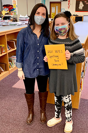picture of teacher standing next to spelling bee champion who is holding a certificate. Both are wearing masks and looking at the camera.