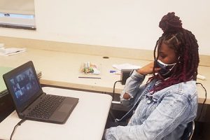 picture of student seated at desk with laptop open to Zoom presentation with reporter
