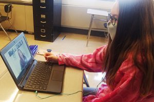 picture of student seated at desk with laptop open to Zoom presentation with reporter