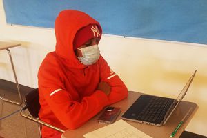 picture of student seated at desk with laptop open to Zoom presentation with reporter