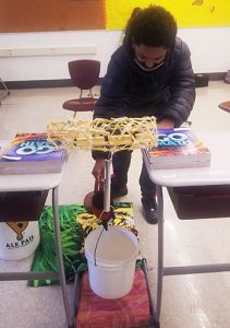 picture of student pouring water from pitcher into bucket to test the strength of a pasta bridge suspended between two desks
