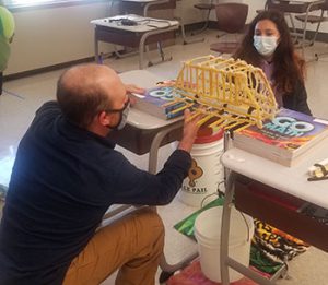 picture of teacher and student checking the strength of a pasta bridge suspended between two desks 