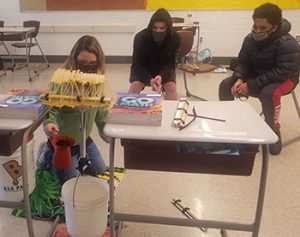 picture of student pouring water from pitcher into bucket to test the strength of a pasta bridge suspended between two desks as other students look on