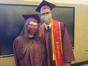 picture of two graduates wearing face masks standing near dressed in caps and gowns