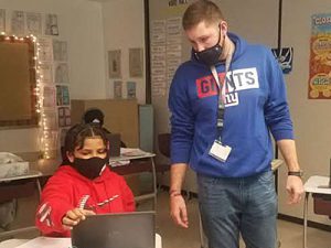 picture of teacher standing next to student seated at desk with laptop open