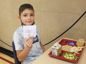 picture of student seated at desk in cafeteria holding up special lunch ticket