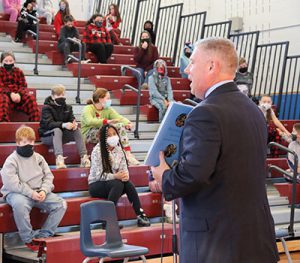 picture of Assemblyman John McDonald holding open a book and reading to students seated in bleachers in school gym.