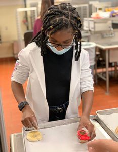 picture of student sprinkling toppings onto rolled bagel dough on a baking sheet