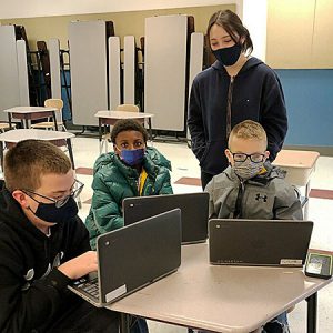 High school senior supports three younger students who are seated at a table working on laptops in the elementary school cafeteria 