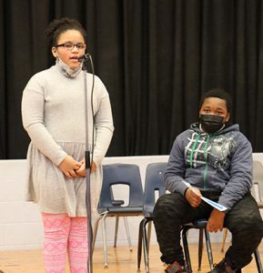 picture of student standing at microphone while another student seated behind watching