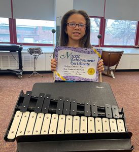 picture of grade 4 Band Student of the Month for February 2022 standing behind musical instrument holding achievement certificate