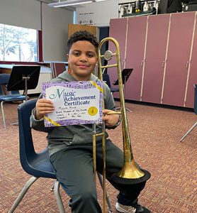 picture of grade 5 Band Student of the Month for February 2022 seated with trombone and holding up achievement certificate