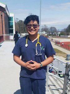 picture of student standing outside school dressed in medical scrubs with hands folded and stethoscope looped around his neck
