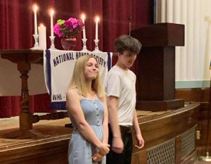picture of two National Honor Society inductees standing together in front of stage with National Honor Society banner and candles representing character, leadership, service and scholarship