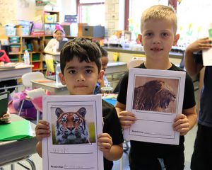 picture of two students standing together looking at camera and smiling. One holds a picture of a tiger on the cover sheet of a class project. The other holds a picture of al lion on the cover sheet of a class project.