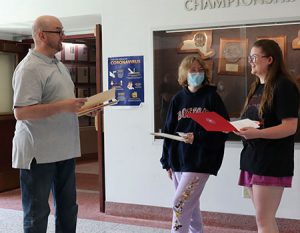 picture of science department director handing folders to the two scholarship winners in the  lobby of the high school building.