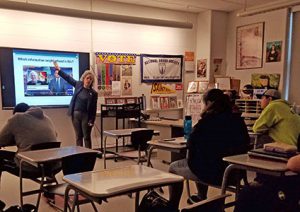 Guest speaker stands in front of classroom facing toward students and pointing at presentation on whiteboard behind her.