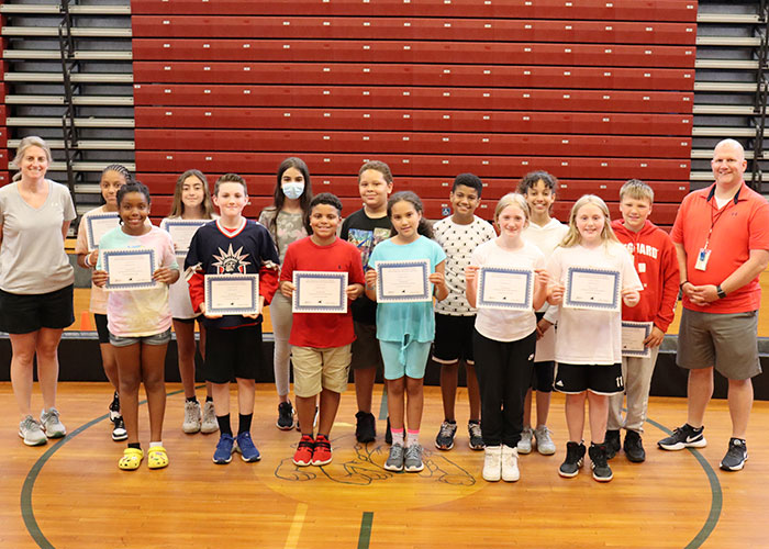 A group of thirteen students standing in the gym holding award certificates with physical education teachers standing to the left and right of the student group.