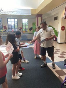 Elks representative hands out certificates to students in the school lobby as art teacher looks on.