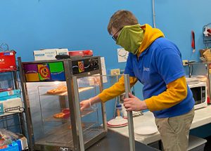 Student with face mask reaching into a heated display oven and grabbing a slice of pizza.