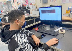 Student with face mask seated at desk looking at computer screen with right hand on mouse.