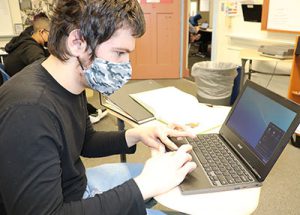 Student with face mask seated at desk typing on keyboard while looking at screen of a laptop computer