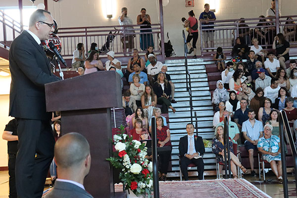 Superintendent at podium speaking into a microphone. Audience visible in the background.