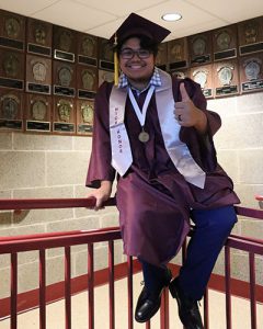 Graduate seated on stair railing looking at camera, smiling with thumbs up.