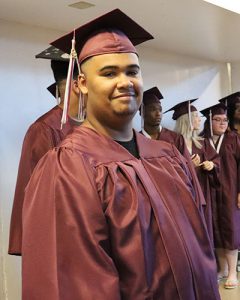 Graduate proceeding in line entering gymnasium looking at camera and smiling.