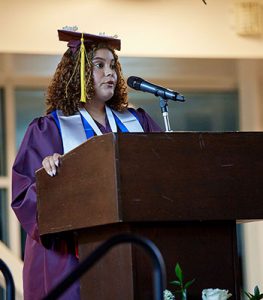 picture of graduation in cap and gown standing at podium speaking into microphone