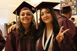 Two graduates stand together smiling at camera. Graduate on right is making the peace sign.