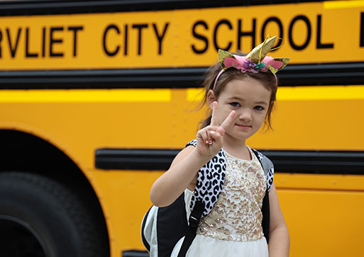 Elementary student holding two fingers up in a peace sign gesture while looking at camera and smiling. Watervliet school bus in the background.