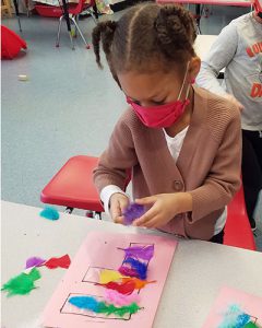 Prekindergarten student seated at desk glues vibrantly colored feathers to pink construction paper that has an outline of the letter  F.