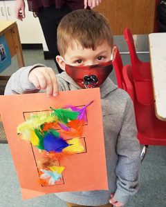 Prekindergarten student looking at camera and holding up a light orange sheet of construction paper that has an outline an outline of the letter F that is filled with vibrantly colored feathers.