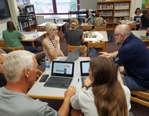 Instructional coach seated at a library table speaking with a teacher. Two other teachers are seated across the table with laptops open.