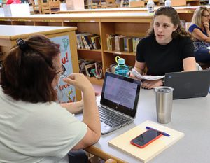 Two teachers seated across from each other at library table, in conversation with laptop computers open.