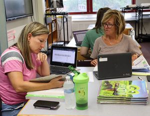 Two teachers seated next to each other with open laptop computers, examining an open textbook.