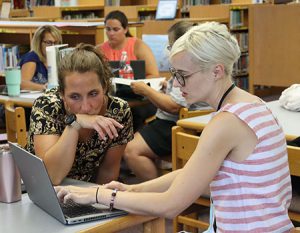 Instructional coach and teacher are seated at a library table speaking and pointing toward screen of open laptop computer.