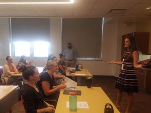 Assistant Superintendent stands at front of room, with arms outstretched speaking to a room full of new teachers and staff.