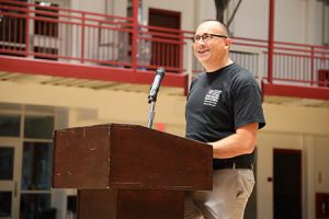 Superintendent of schools dressed in black United in Kindness t-shirt, standing at podium smiling out at audience.