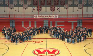 Teachers, staff dressed in United in Kindness t-shirts gather in center of gymnasium floor in the formation of a W.