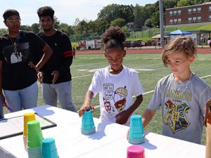 Two students compete in cup stacking game as siblings look on and cheer.