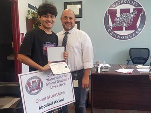 School counselor stands beside the new graduate who is holding high school diploma and Class of 2002 lawn sign. Both are facing the camera and smiling.