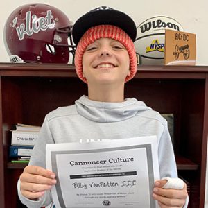 grade 8 student wearing a ball cap and smiling at camera holding Cannoneer Culture certificate for Being Proud