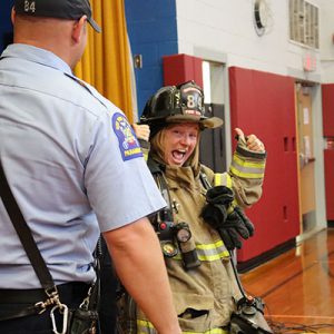 A Watervliet staff member dressed in firefighting gear smiles at camera with thumbs up sign as a firefighter looks on nearby during fire safety assembly for students.