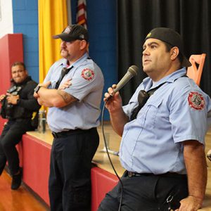 A Watervliet firefighter leans against the stage in the elementary school speaking into a microphone during fire safety assembly for students.